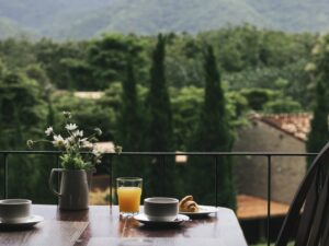 Mesa de café da manhã em varanda: suco, café, croissant e flores, vista para montanhas.