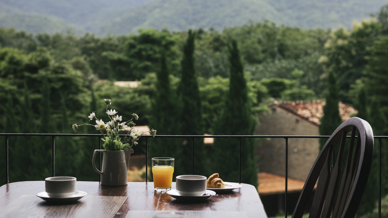Mesa de café da manhã em varanda: suco, café, croissant e flores, vista para montanhas.