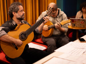Músico toca violão de sete cordas em destaque, com outros integrantes de um grupo regional ao fundo em um palco de madeira. Cena de apresentação de música instrumental brasileira