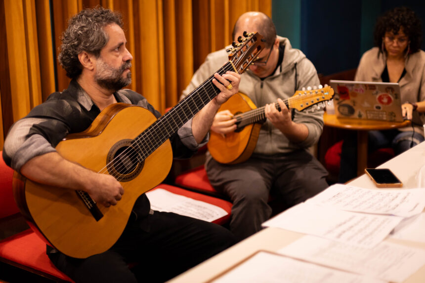 Músico toca violão de sete cordas em destaque, com outros integrantes de um grupo regional ao fundo em um palco de madeira. Cena de apresentação de música instrumental brasileira