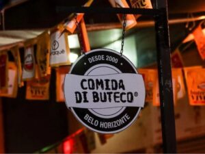 Sinal pendurado preto e branco com "COMIDA DI BUTECO DESDE 2000 BELO HORIZONTE" e banners amarelos.