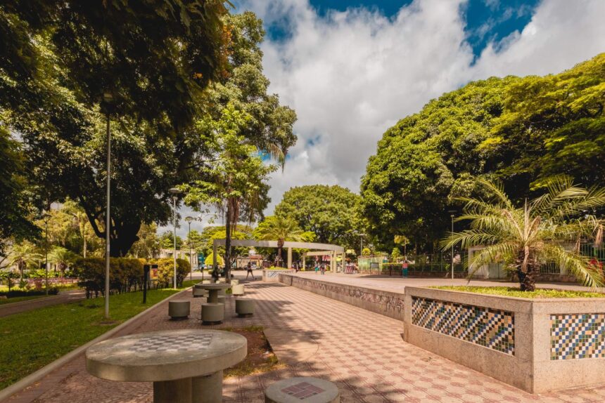 Vista de uma praça arborizada com mesas de xadrez de concreto, pavimento decorado e jardim