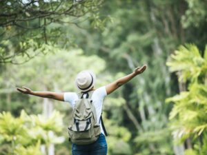 Pessoa de costas, braços abertos, com mochila e chapéu em paisagem natural verde e exuberante.