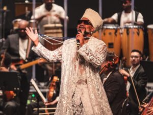 Carlinhos Brown canta em palco, usando roupa branca e turbante, com músicos de orquestra ao fundo.