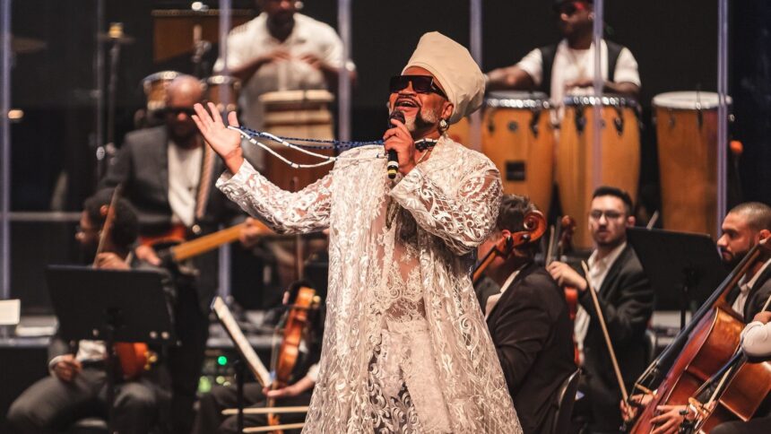 Carlinhos Brown canta em palco, usando roupa branca e turbante, com músicos de orquestra ao fundo.