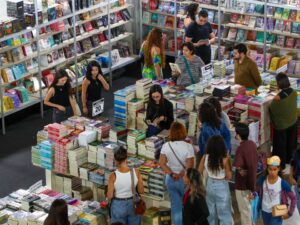 Pessoas em feira de livros, olhando prateleiras e pilhas de volumes. Cartazes com '20', 'Literatura', 'Oferta' visíveis.