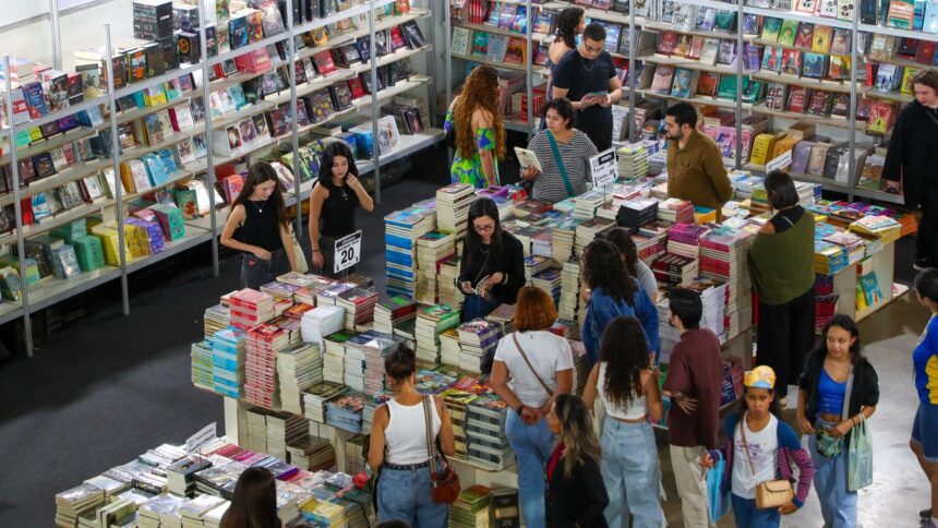 Pessoas em feira de livros, olhando prateleiras e pilhas de volumes. Cartazes com '20', 'Literatura', 'Oferta' visíveis.