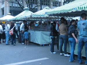 Pessoas em feira de rua com barracas listradas verdes e brancas movimentam o comércio.