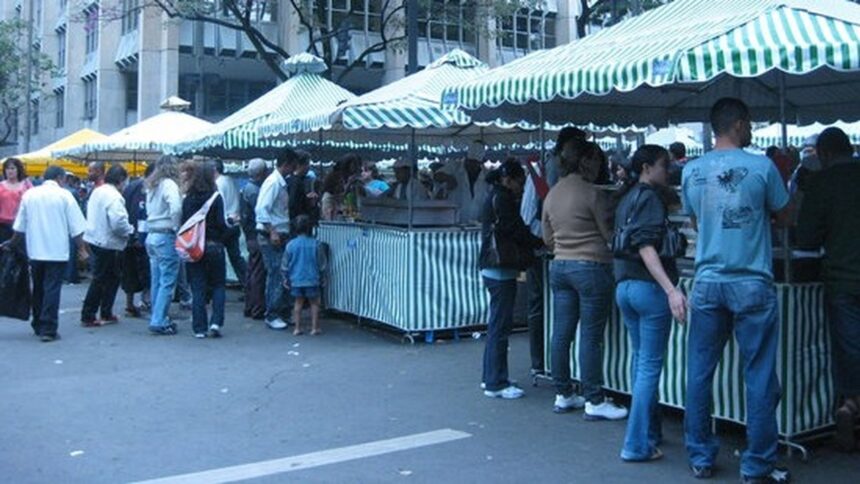 Pessoas em feira de rua com barracas listradas verdes e brancas movimentam o comércio.