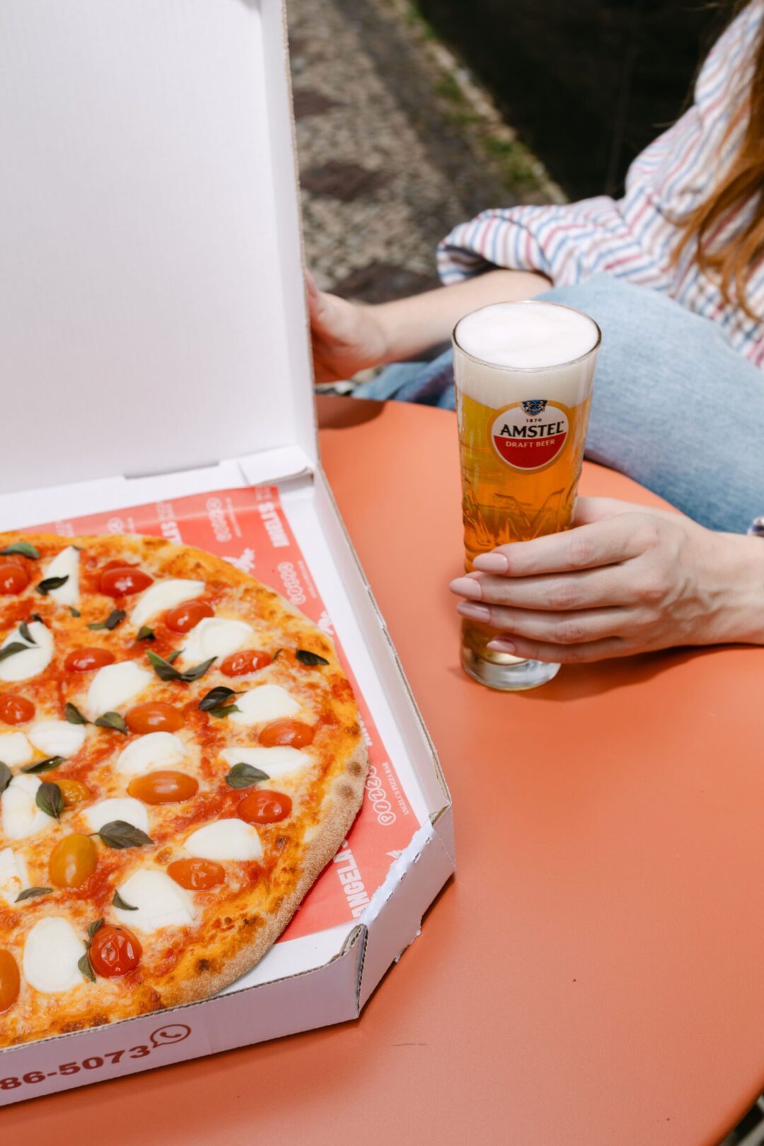 Mão segura copo de Amstel Draft Beer ao lado de pizza margherita em caixa. - (foto: Barbara Dutra)