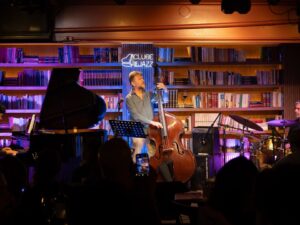 Três músicos de jazz tocam piano, contrabaixo e bateria em palco de clube, com neon 'CLUBE DEJAZZ'.