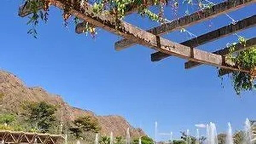 Céu azul sobre pérgola de madeira com folhagem verde e montanhas, fontes de água em parque.