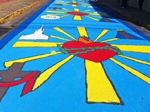 Tapete azul com símbolos religiosos: Coração de Jesus, cruz, igreja e 'JHS' em rua.