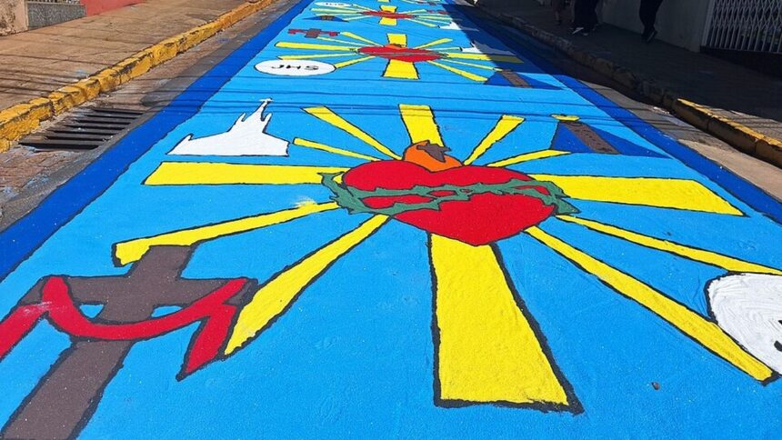 Tapete azul com símbolos religiosos: Coração de Jesus, cruz, igreja e 'JHS' em rua.
