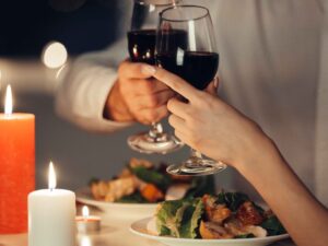 Mãos de um casal brindando com taças de vinho tinto, velas acesas e jantar à mesa.