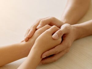 Close-up de duas mãos adultas sobrepostas, uma segurando e outra sendo segurada, em fundo claro.