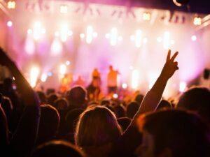 Pessoas em um festival com mãos levantadas, de costas para o palco iluminado.