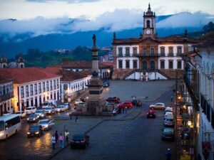 Praça Tiradentes em Ouro Preto ao anoitecer, com carros, pessoas, monumento e edifício colonial.