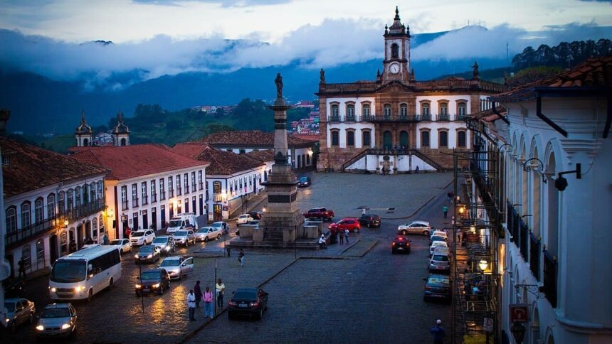 Praça Tiradentes em Ouro Preto ao anoitecer, com carros, pessoas, monumento e edifício colonial.