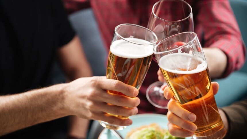 Três pessoas brindam com dois copos de cerveja e uma taça de vinho em um bar.