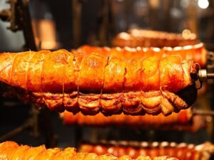 Vários pedaços de carne suína dourada, amarrados com barbante, girando em espetos de rotisserie.