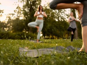 Três mulheres praticam yoga em postura da árvore sobre tapetes na grama de um parque.