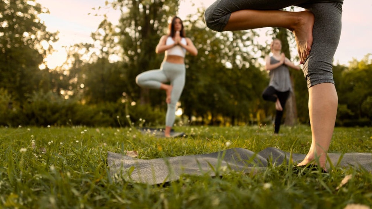 Três mulheres praticam yoga em postura da árvore sobre tapetes na grama de um parque.