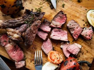 Corte de carne grelhada na brasa e fatiada em tábua de madeira com legumes e temperos.