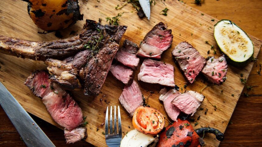 Corte de carne grelhada na brasa e fatiada em tábua de madeira com legumes e temperos.