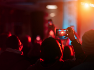 Pessoa filmando show com smartphone em evento lotado, luzes vibrantes no palco.