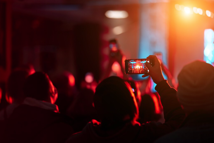 Pessoa filmando show com smartphone em evento lotado, luzes vibrantes no palco.