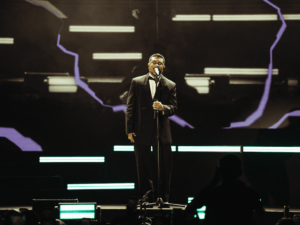 Cantor Xamã de terno preto e gravata borboleta no palco com microfone em show.