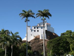 Santuário Convento da Penha branco no alto de penhasco rochoso, com palmeiras e céu azul