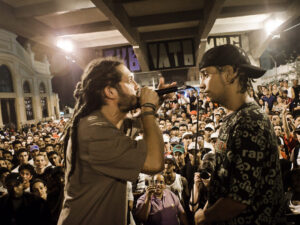Homem com dreadlocks canta em microfone para outro homem em meio à multidão noturna.