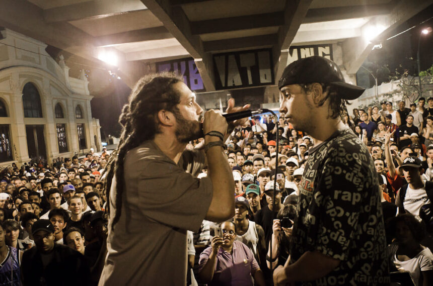 Homem com dreadlocks canta em microfone para outro homem em meio à multidão noturna.