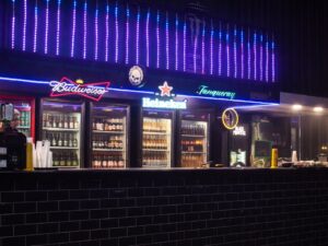 Balcão de bar de tijolos pretos com geladeiras iluminadas, rótulos Budweiser, Heineken e Tanqueray.
