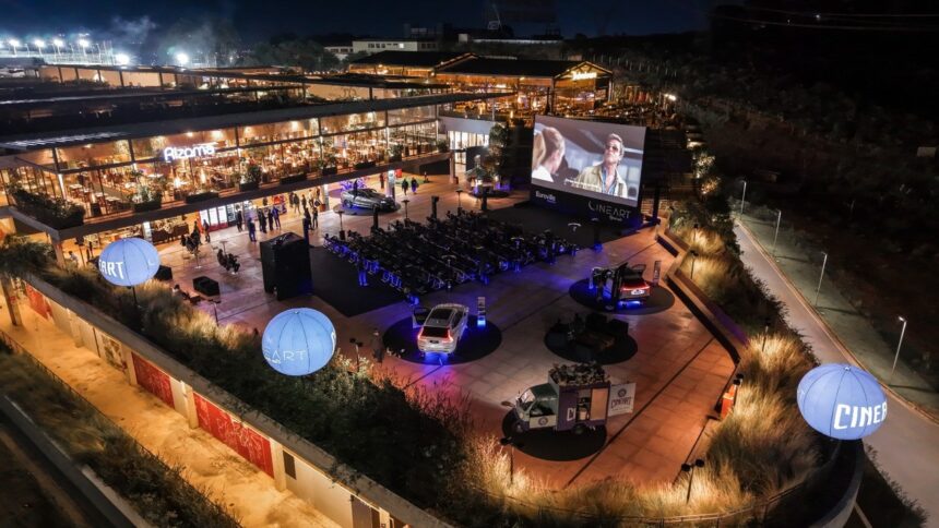 Vista aérea noturna de cinema ao ar livre com tela, cadeiras e prédios iluminados.