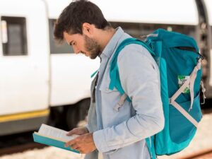 Homem com mochila azul lendo um livro em uma plataforma de trem.