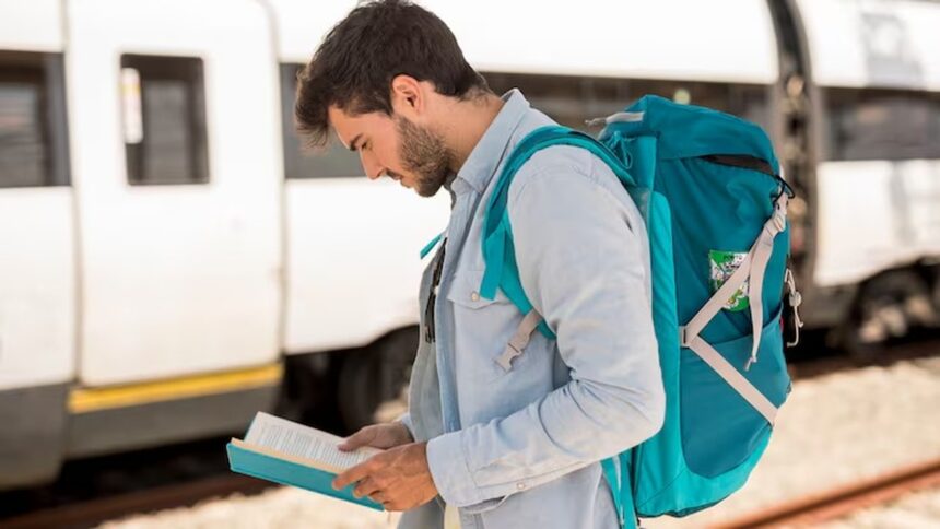 Homem com mochila azul lendo um livro em uma plataforma de trem.