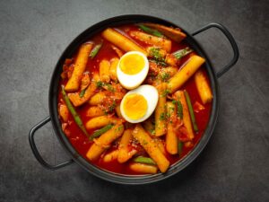 Tteokbokki em molho vermelho com ovos, vegetais verdes e gergelim em panela preta.
