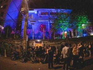 Fachada de centro cultural iluminada em azul e verde, com pessoas e motos na rua à noite.