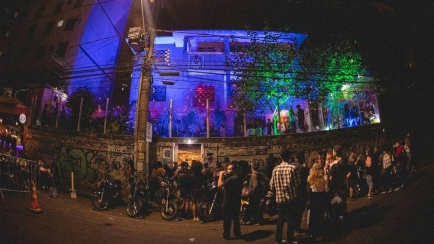 Fachada de centro cultural iluminada em azul e verde, com pessoas e motos na rua à noite.