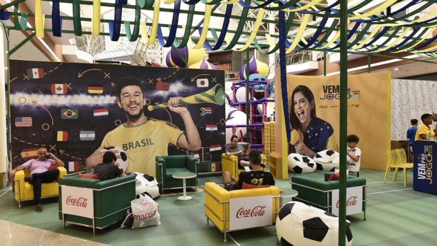 Pessoas em espaço temático de futebol “VEM PRO JOGO”, com banners e logos Coca-Cola, Panini.