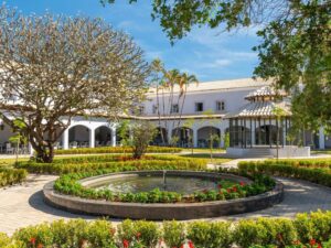 Jardim do hotel com fonte circular, árvores, gazebo e prédio branco histórico.
