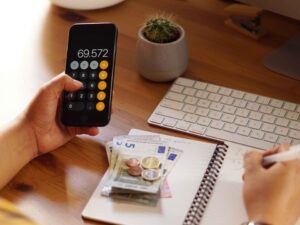 Mãos usam calculadora em celular com '69.572', anotam em caderno, com euros e teclado.