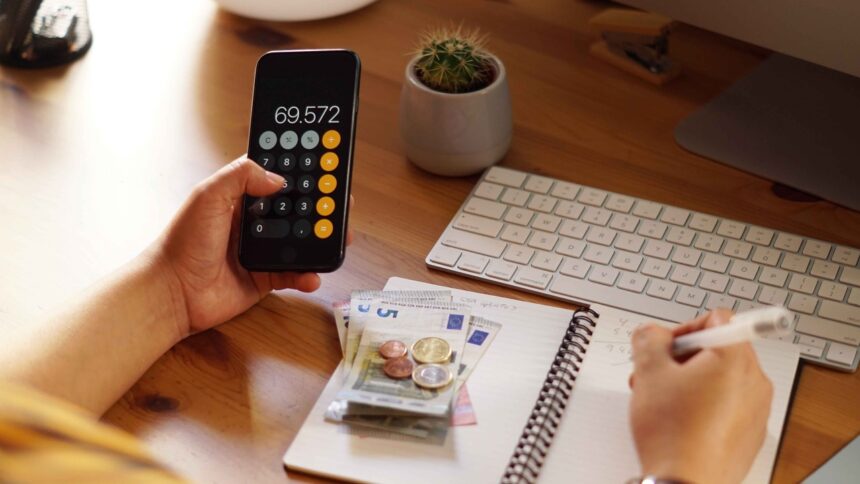 Mãos usam calculadora em celular com '69.572', anotam em caderno, com euros e teclado.