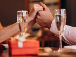 Mãos de casal segurando-se em jantar romântico, com taças de champanhe, presente e rosa.