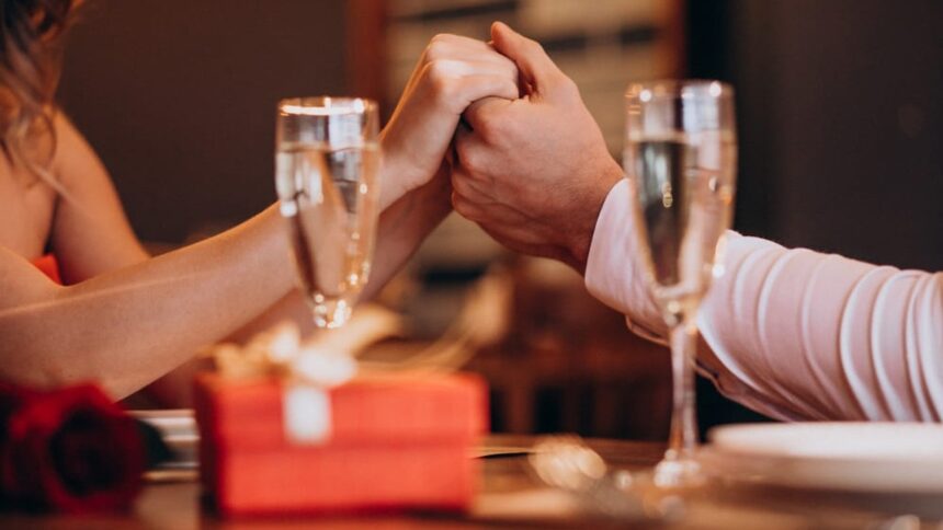 Mãos de casal segurando-se em jantar romântico, com taças de champanhe, presente e rosa.