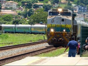 Locomotiva 1290 chegando à estação, pessoas na plataforma, outro trem e cidade ao fundo.