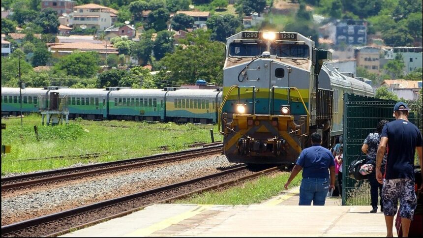 Locomotiva 1290 chegando à estação, pessoas na plataforma, outro trem e cidade ao fundo.